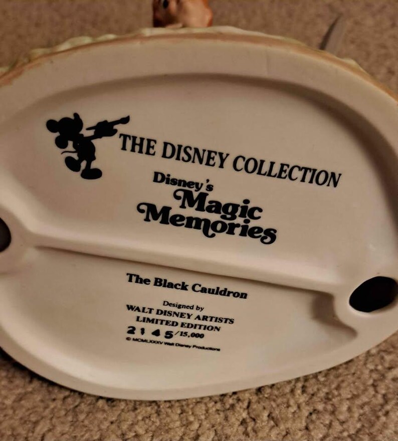 May include: A white ceramic base of a Disney figurine, featuring the text "THE DISNEY COLLECTION" and "Disney's Magic Memories." The base is marked "The Black Cauldron" and "LIMITED EDITION 2145/15,000."