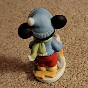 May include: A ceramic figurine of Mickey Mouse dressed in winter clothing, wearing a blue hat with a white pom-pom, a green and white scarf, and red trousers. He is holding ski poles and standing on skis.