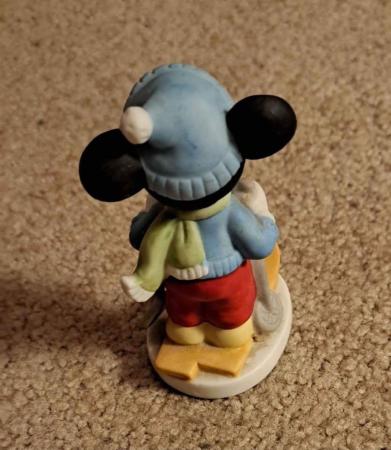 May include: A ceramic figurine of Mickey Mouse dressed in winter clothing, wearing a blue hat with a white pom-pom, a green and white scarf, and red trousers. He is holding ski poles and standing on skis.