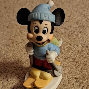 May include: A ceramic figurine of Mickey Mouse wearing a blue hat and red trousers, skiing down a snowy slope.