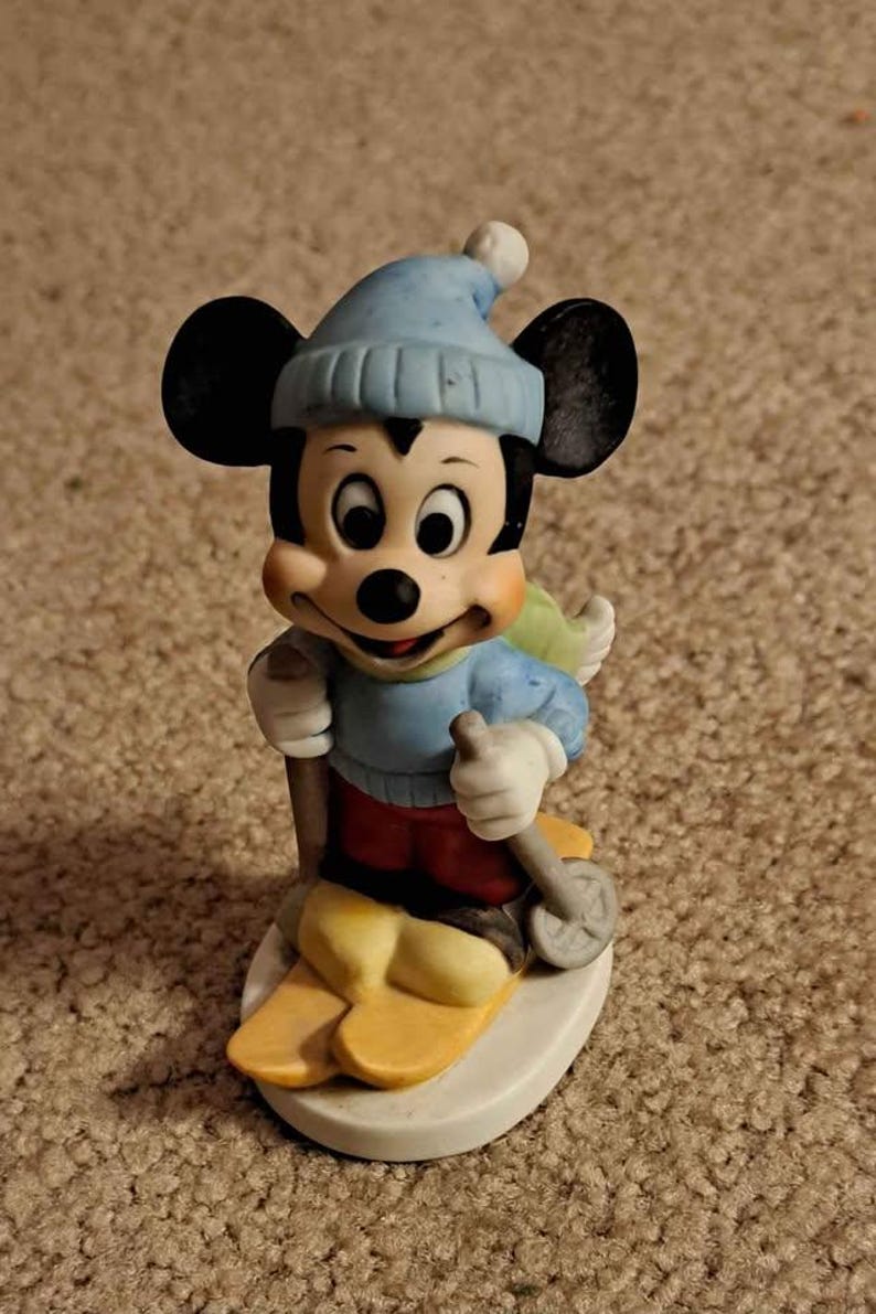 May include: A ceramic figurine of Mickey Mouse wearing a blue hat and red trousers, skiing down a snowy slope.