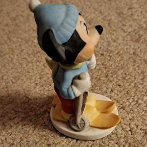 May include: A ceramic figurine of Mickey Mouse wearing a blue hat and red trousers, skiing on yellow skis with a brown ski pole.