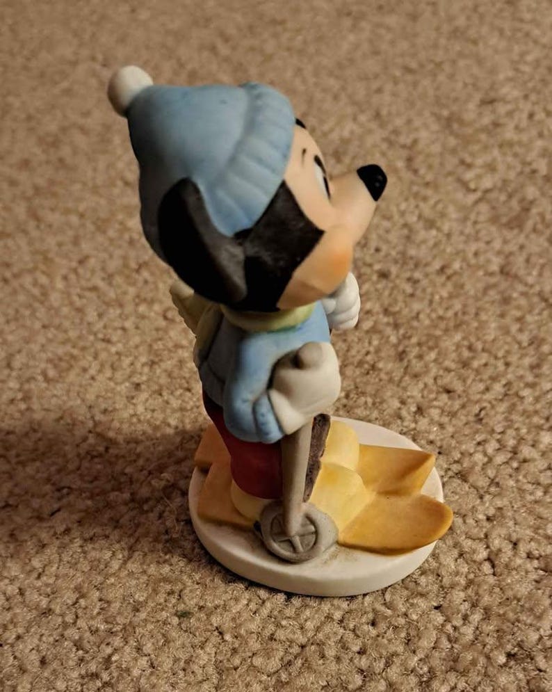 May include: A ceramic figurine of Mickey Mouse wearing a blue hat and red trousers, skiing on yellow skis with a brown ski pole.