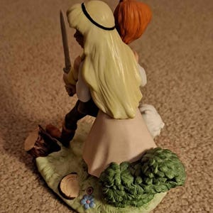 May include: A ceramic figurine depicting two figures in a fairytale scene. One figure, with long blonde hair and a black headband, holds a sword. The other figure has red hair. The base features a grassy area with a tree stump, flowers, and green shrubbery.