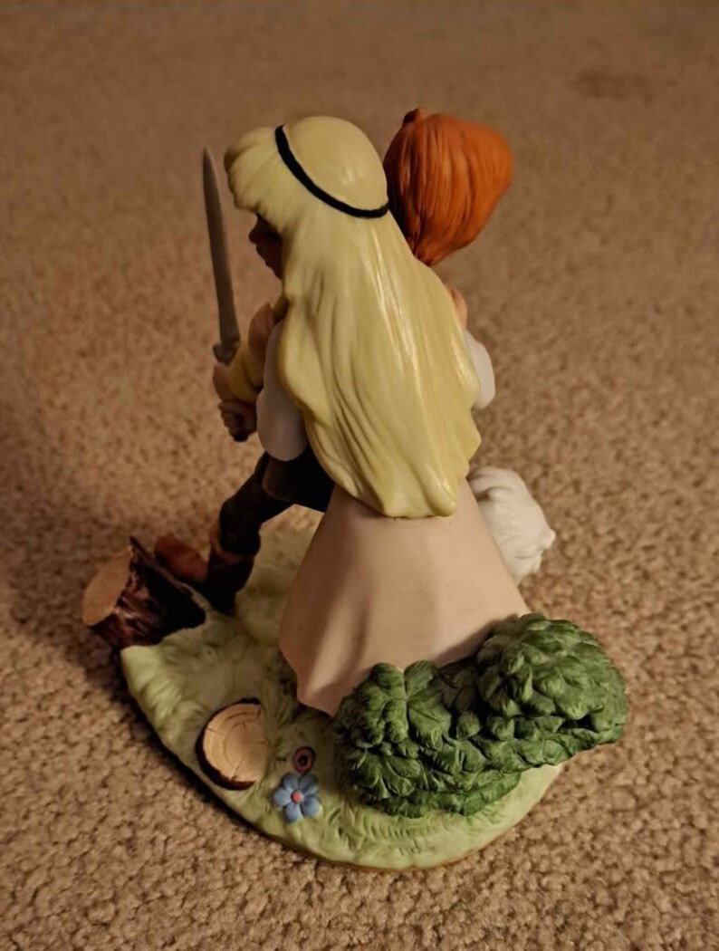 May include: A ceramic figurine depicting two figures in a fairytale scene. One figure, with long blonde hair and a black headband, holds a sword. The other figure has red hair. The base features a grassy area with a tree stump, flowers, and green shrubbery.