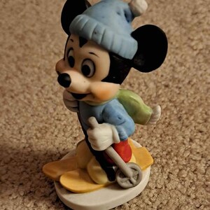 May include: A ceramic figurine of Mickey Mouse wearing a blue hat and scarf, skiing down a snowy slope. He is holding ski poles and has a determined expression on his face.