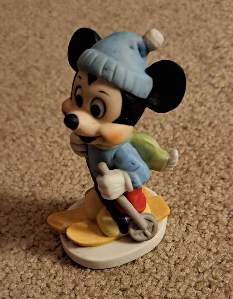 May include: A ceramic figurine of Mickey Mouse wearing a blue hat and scarf, skiing down a snowy slope. He is holding ski poles and has a determined expression on his face.