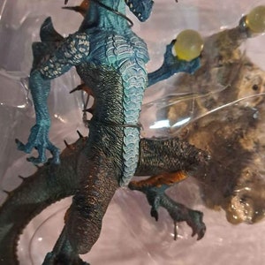 May include: A blue and green dragon figurine with orange wings and a green orb in its hand. The dragon is posed in a sitting position with its tail curled around its body. The figurine is part of a "Sorcerer's Dragon" collection.