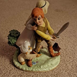May include: A ceramic figurine depicting a young boy with red hair holding a sword, accompanied by a sheep and a woman. The figures are on a green base with a tree and flowers. The boy wears a yellow shirt and brown vest.
