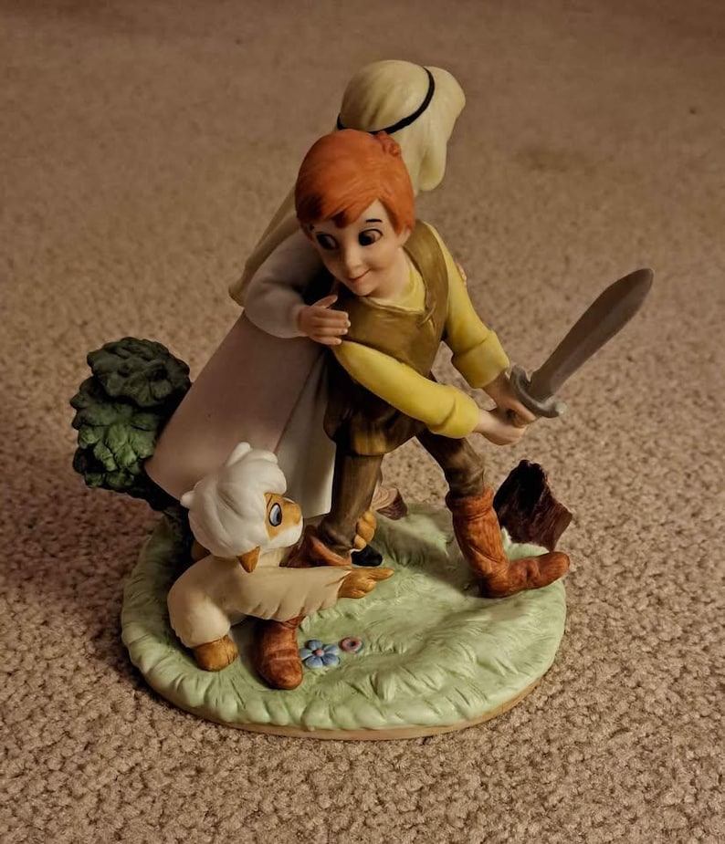 May include: A ceramic figurine depicting a young boy with red hair holding a sword, accompanied by a sheep and a woman. The figures are on a green base with a tree and flowers. The boy wears a yellow shirt and brown vest.