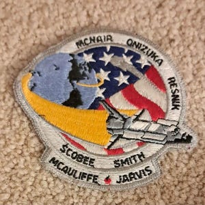 May include: Embroidered patch featuring a space shuttle design with the American flag and a depiction of Earth. The patch includes the names of the Challenger crew: McNair, Onizuka, Resnik, Scobee, McAuliffe, Smith, and Jarvis.