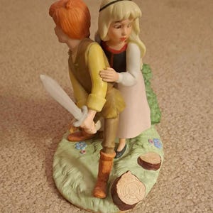 May include: A porcelain figurine depicting a young man with red hair holding a sword, and a young woman with blonde hair. The figures are on a green base with flowers and tree stumps. The woman is wearing a pink dress.