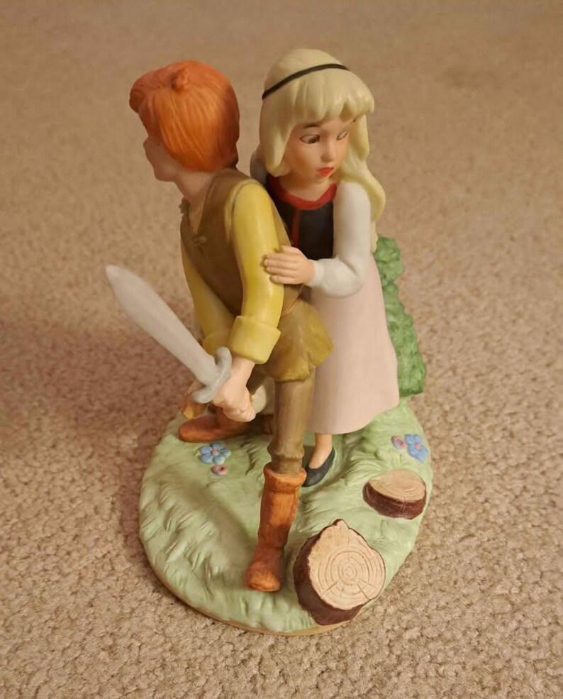 May include: A porcelain figurine depicting a young man with red hair holding a sword, and a young woman with blonde hair. The figures are on a green base with flowers and tree stumps. The woman is wearing a pink dress.