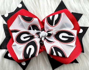 May include: A red, white, and black bow with a University of Georgia logo. The bow is made of ribbon and has a polka dot pattern on the back.