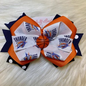 Puede incluir: Un lazo para el pelo blanco, naranja y azul marino con un dije de baloncesto en el centro. El lazo tiene el logotipo de los Oklahoma City Thunder impreso en él.