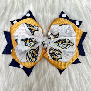 Puede incluir: Un lazo blanco y dorado con un borde de lunares azul marino y blancos. El lazo presenta un patrón repetido de un logotipo blanco y azul.