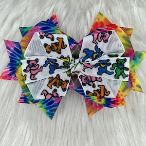 May include: A colorful tie-dye hair bow with a white center featuring a pattern of dancing bears. The bow is made of two layers of ribbon, with the tie-dye pattern on the outer layer and the bear pattern on the inner layer.