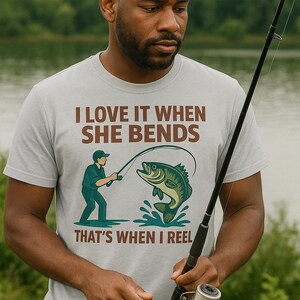 May include: A light gray t-shirt with the text "I LOVE IT WHEN SHE BENDS, THAT'S WHEN I REEL" and a graphic of a fisherman reeling in a fish. The person in the image is holding a fishing rod.