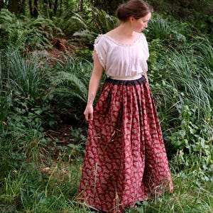 May include: A woman wearing a white blouse with lace trim and a long, flowing red skirt with a floral pattern. The skirt has a black waistband. The scene is set outdoors, surrounded by lush green foliage and trees.