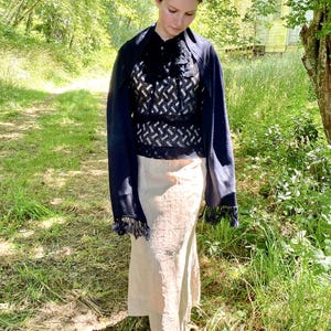 Puede incluir: Una mujer con un top estampado en blanco y negro, un chal azul marino con flecos y una falda beige. El atuendo se usa al aire libre en una zona herbosa con &aacute;rboles y un edificio al fondo.