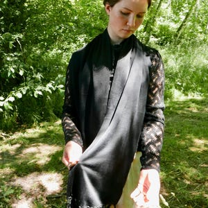 Puede incluir: Una bufanda negra con borde de flecos est&aacute; drapeada alrededor del cuello. La bufanda est&aacute; hecha de una tela suave y fluida. La persona lleva una blusa negra con mangas de encaje. El fondo es un exuberante bosque verde.