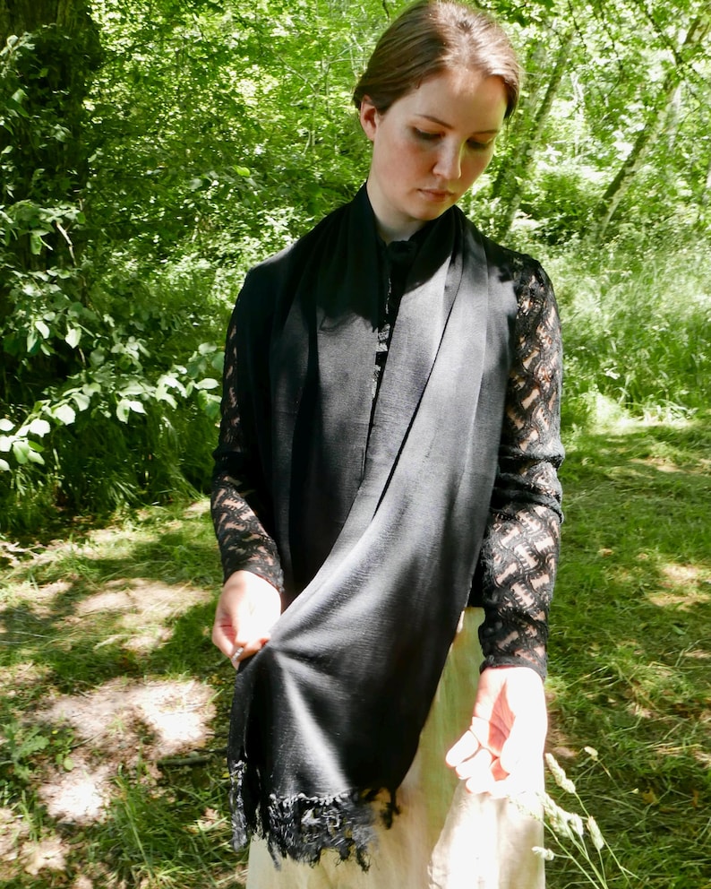 Puede incluir: Una bufanda negra con borde de flecos est&aacute; drapeada alrededor del cuello. La bufanda est&aacute; hecha de una tela suave y fluida. La persona lleva una blusa negra con mangas de encaje. El fondo es un exuberante bosque verde.