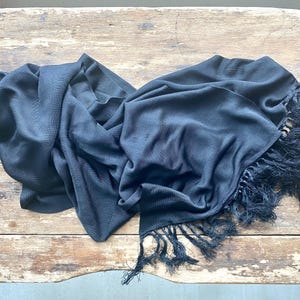 Puede incluir: Una bufanda azul marino oscuro con una textura sedosa y bordes con flecos. La bufanda est&aacute; drapeada sobre una superficie de madera r&uacute;stica, mostrando su tejido fluido y su color oscuro. Los bordes con flecos a&ntilde;aden un elemento decorativo.