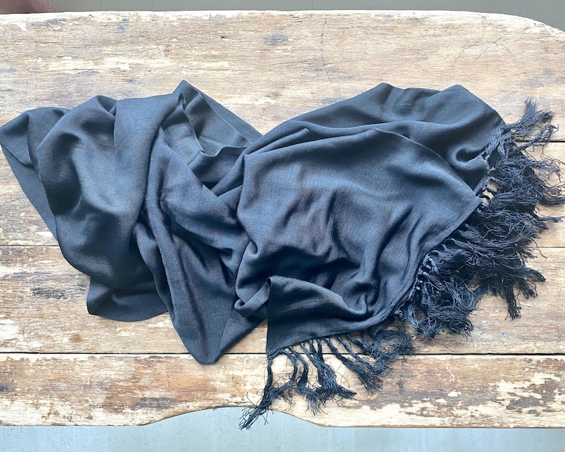 Puede incluir: Una bufanda azul marino oscuro con una textura sedosa y bordes con flecos. La bufanda est&aacute; drapeada sobre una superficie de madera r&uacute;stica, mostrando su tejido fluido y su color oscuro. Los bordes con flecos a&ntilde;aden un elemento decorativo.