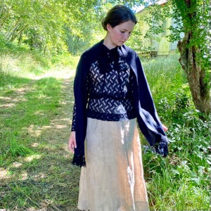 Puede incluir: Una mujer con un atuendo vintage al aire libre. El conjunto incluye un chal azul marino, una blusa con estampado en blanco y negro y una falda beige. La escena se desarrolla en un camino cubierto de hierba con &aacute;rboles y un edificio al fondo.