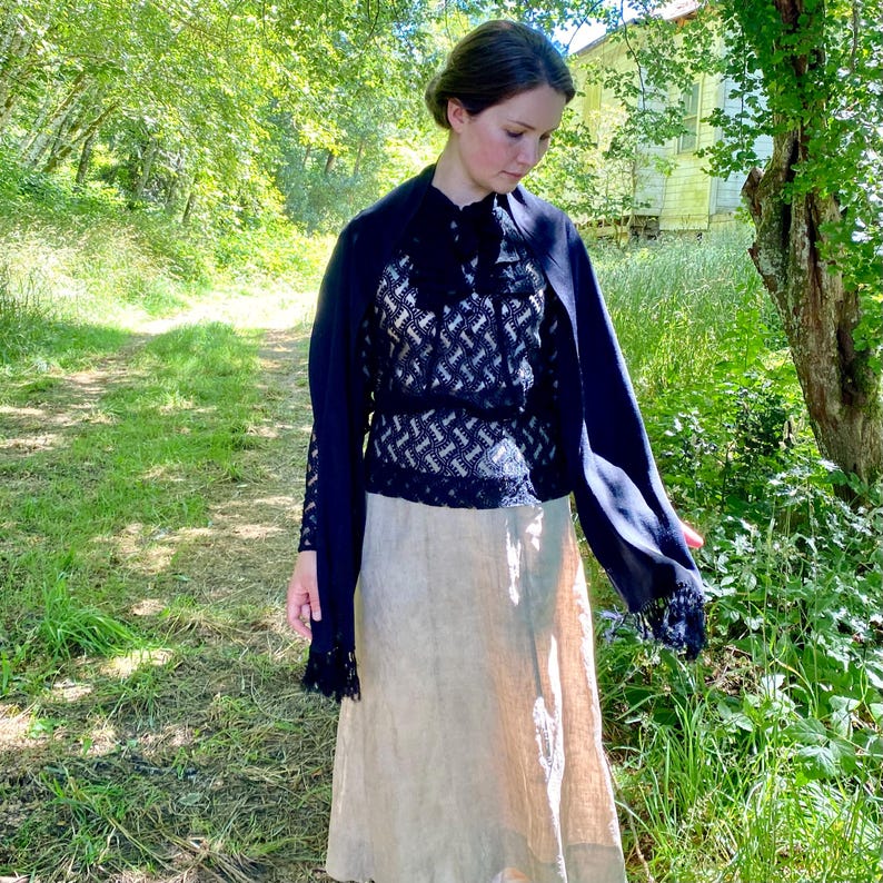 Puede incluir: Una mujer con un atuendo vintage al aire libre. El conjunto incluye un chal azul marino, una blusa con estampado en blanco y negro y una falda beige. La escena se desarrolla en un camino cubierto de hierba con &aacute;rboles y un edificio al fondo.