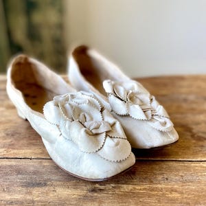 Op de afbeelding: Een paar witte satijnen pantoffels met een grote witte bloemversiering op de teen.