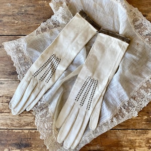 May include: A pair of vintage cream-colored gloves with decorative black stitching and a dark brown cuff. The gloves are laid on a white lace-trimmed cloth, set against a rustic wooden surface. The gloves have a classic, elegant design.