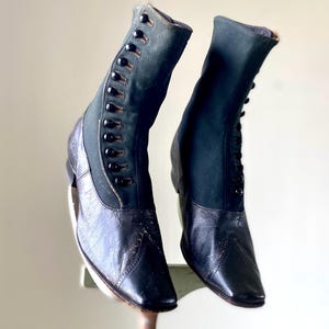 Puede incluir: Un par de botas de cuero negro con una parte superior de lona verde oscuro. Las botas tienen una punta puntiaguda y un cierre de botón en la parte delantera.