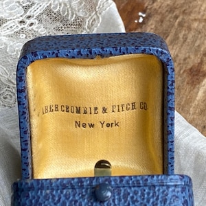 Op de afbeelding: Open blau lederen Abercrombie & Fitch Co. sieradendoosje met een gouden satijnen binnenkant. De doos is open en toont de merknaam en de stad New York.
