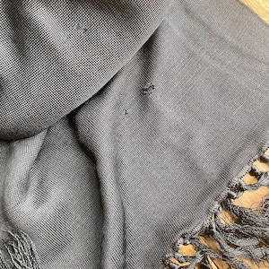 Puede incluir: Primer plano de una bufanda gris oscuro con un tejido texturizado y flecos. La bufanda est&aacute; drapeada, mostrando la textura de la tela y los flecos a lo largo del borde. La bufanda parece estar hecha de un material suave.