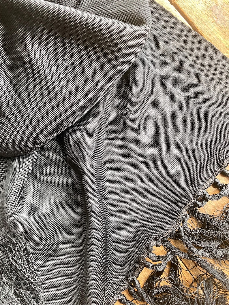 Puede incluir: Primer plano de una bufanda gris oscuro con un tejido texturizado y flecos. La bufanda est&aacute; drapeada, mostrando la textura de la tela y los flecos a lo largo del borde. La bufanda parece estar hecha de un material suave.