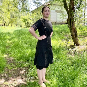 May include: A black dress with a white button-up front and a white collar. The dress has a short sleeve and a skirt that falls to the knees. The dress is worn by a person standing in a grassy field.