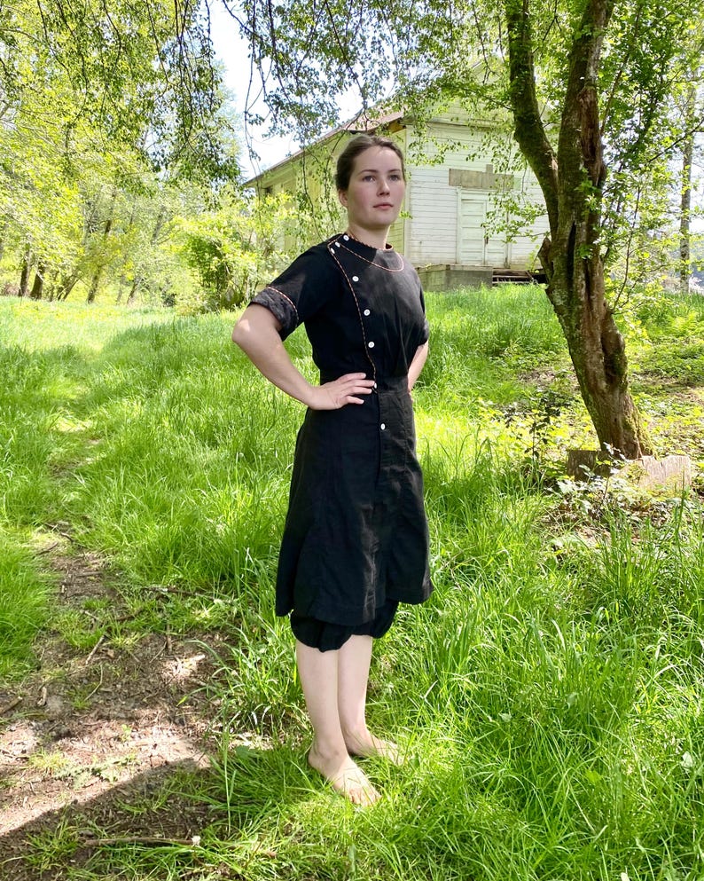 May include: A black dress with a white button-up front and a white collar. The dress has a short sleeve and a skirt that falls to the knees. The dress is worn by a person standing in a grassy field.