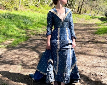 May include: A long, blue dress with a floral pattern and white lace trim. The dress has long sleeves and a ruffled skirt. The person is standing on a dirt path in a forest setting.
