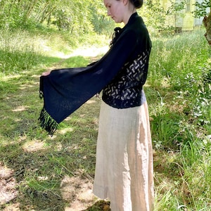 Puede incluir: Un chal negro con mangas largas y acampanadas y ribete de flecos est&aacute; drapeado sobre una falda beige. El chal tiene una espalda con un patr&oacute;n de tejido abierto. El atuendo se usa al aire libre en un entorno natural con luz solar.