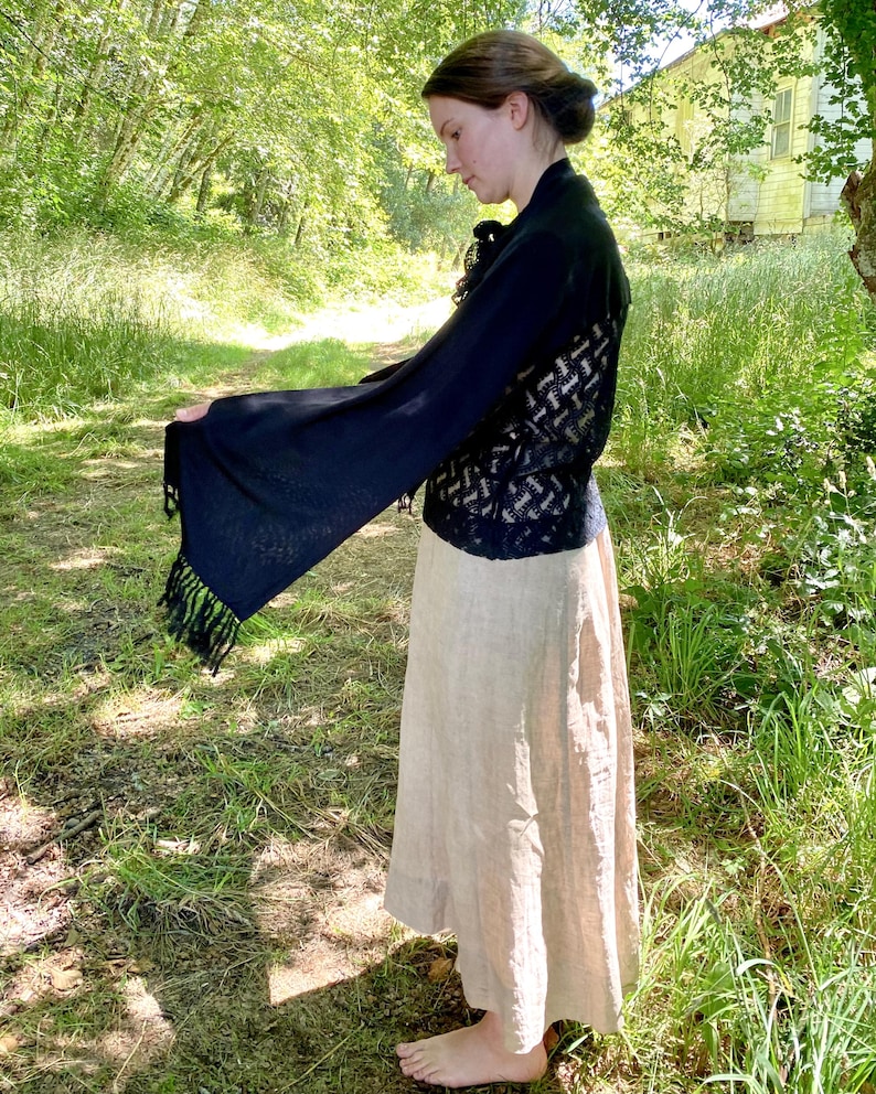 Puede incluir: Un chal negro con mangas largas y acampanadas y ribete de flecos est&aacute; drapeado sobre una falda beige. El chal tiene una espalda con un patr&oacute;n de tejido abierto. El atuendo se usa al aire libre en un entorno natural con luz solar.