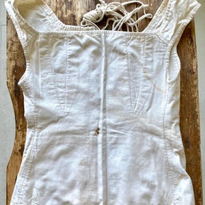 May include: Cream-colored, sleeveless top with a square neckline and shoulder straps. The garment has vertical seams and a drawstring at the back. The fabric appears to be linen or a similar natural fiber.