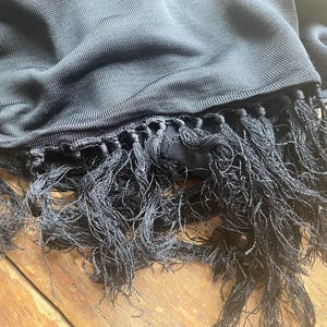 Puede incluir: Primer plano de una bufanda negra con un tejido texturizado y largas borlas anudadas. La bufanda est&aacute; drapeada sobre una superficie de madera, mostrando la suavidad de la tela y los detalles de las borlas.