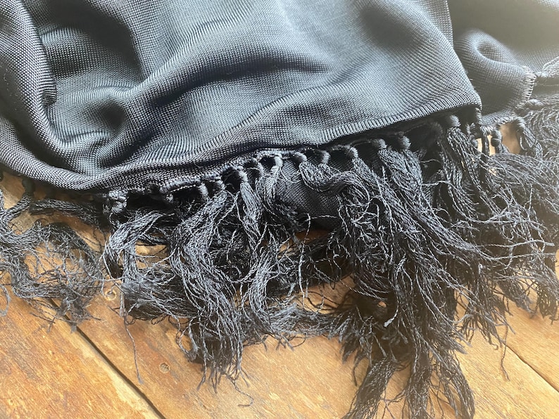 Puede incluir: Primer plano de una bufanda negra con un tejido texturizado y largas borlas anudadas. La bufanda est&aacute; drapeada sobre una superficie de madera, mostrando la suavidad de la tela y los detalles de las borlas.