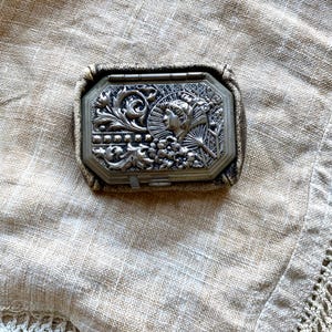 May include: An antique silver-toned compact with an ornate, raised design featuring a woman's profile, floral elements, and a fan. The compact is rectangular with an octagonal lid and a dark, textured border. The background is a neutral linen fabric.
