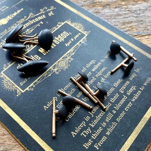 May include: A collection of black cufflinks with gold-colored stems, resting on a vintage black card with gold trim. The card features ornate text and a decorative border. The cufflinks have a rounded, oval shape.