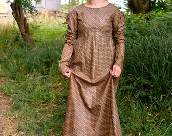 Puede incluir: Un vestido largo marrón con un patrón de puntos repetidos. El vestido tiene mangas largas con hombros abullonados y cintura alta. La mujer está de pie al aire libre en una zona cubierta de hierba.