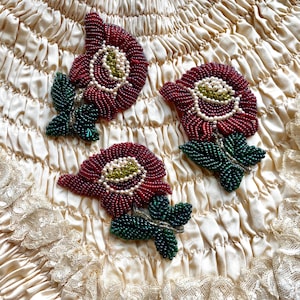 Könnte beinhalten: Drei gebastelte Blumenbroschen mit roten Blütenblättern, weißen Perlenzentren und grünen Perlenblättern. Die Broschen befinden sich auf einem cremefarbenen, strukturierten Stoff mit Spitzensaum. Die Blumen haben ein detailliertes, handgefertigtes Aussehen.