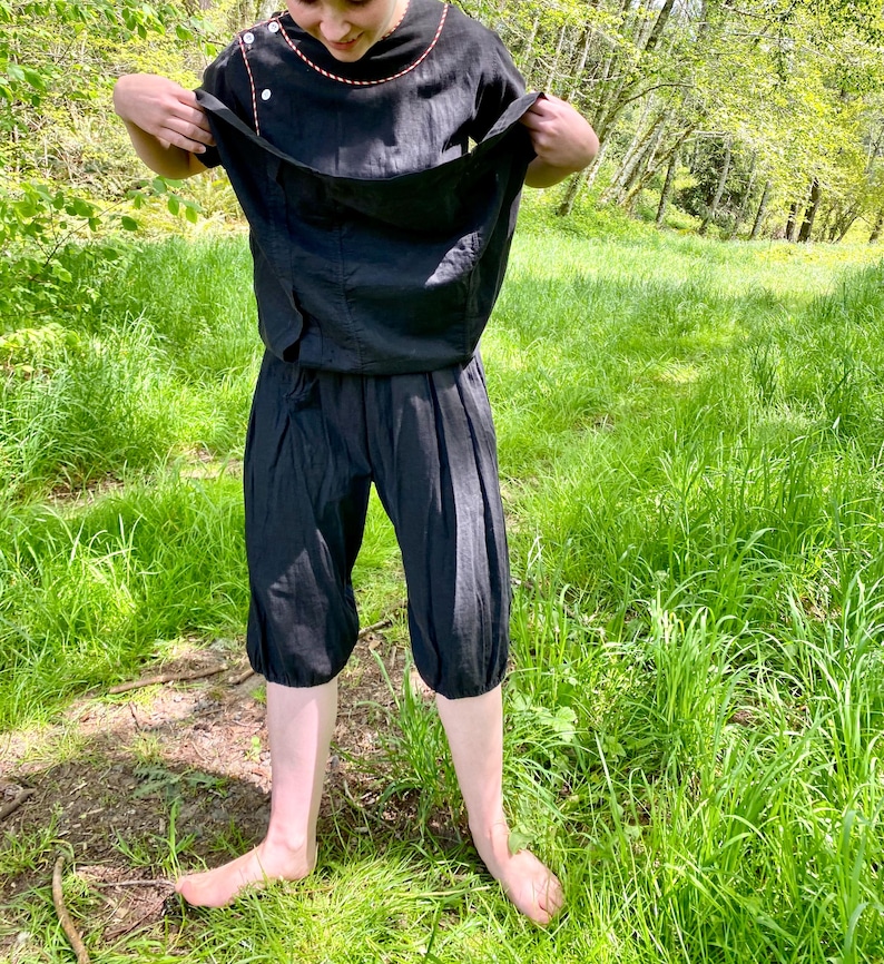 May include: A black two-piece outfit with a short-sleeved top and pants. The top has a red and white trim around the neckline and a button on the front. The pants are loose-fitting and have a gathered look at the bottom.