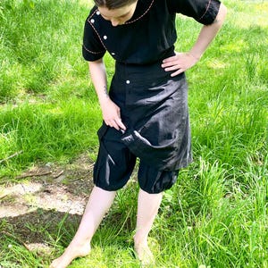 May include: A black dress with a contrasting orange stitch detail. The dress has a button-up front and a gathered skirt. The model is wearing the dress with a pair of black bloomers.